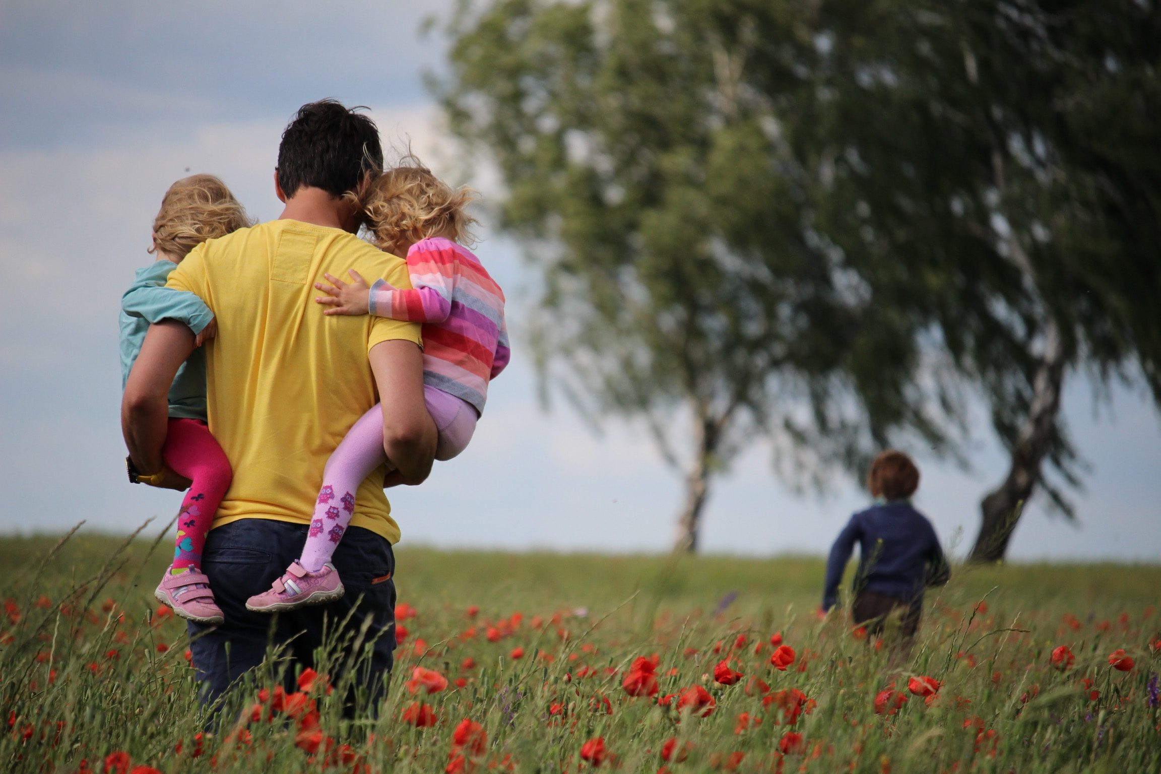A father carries his two children in a field of red flowers, with another child running ahead to a nearby tree.